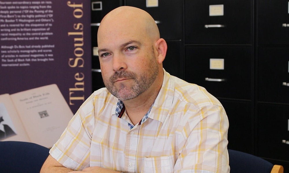 A man with a bald head and trimmed beard sits at a desk, wearing a plaid shirt. Filing cabinets and a poster with text are visible in the background. 