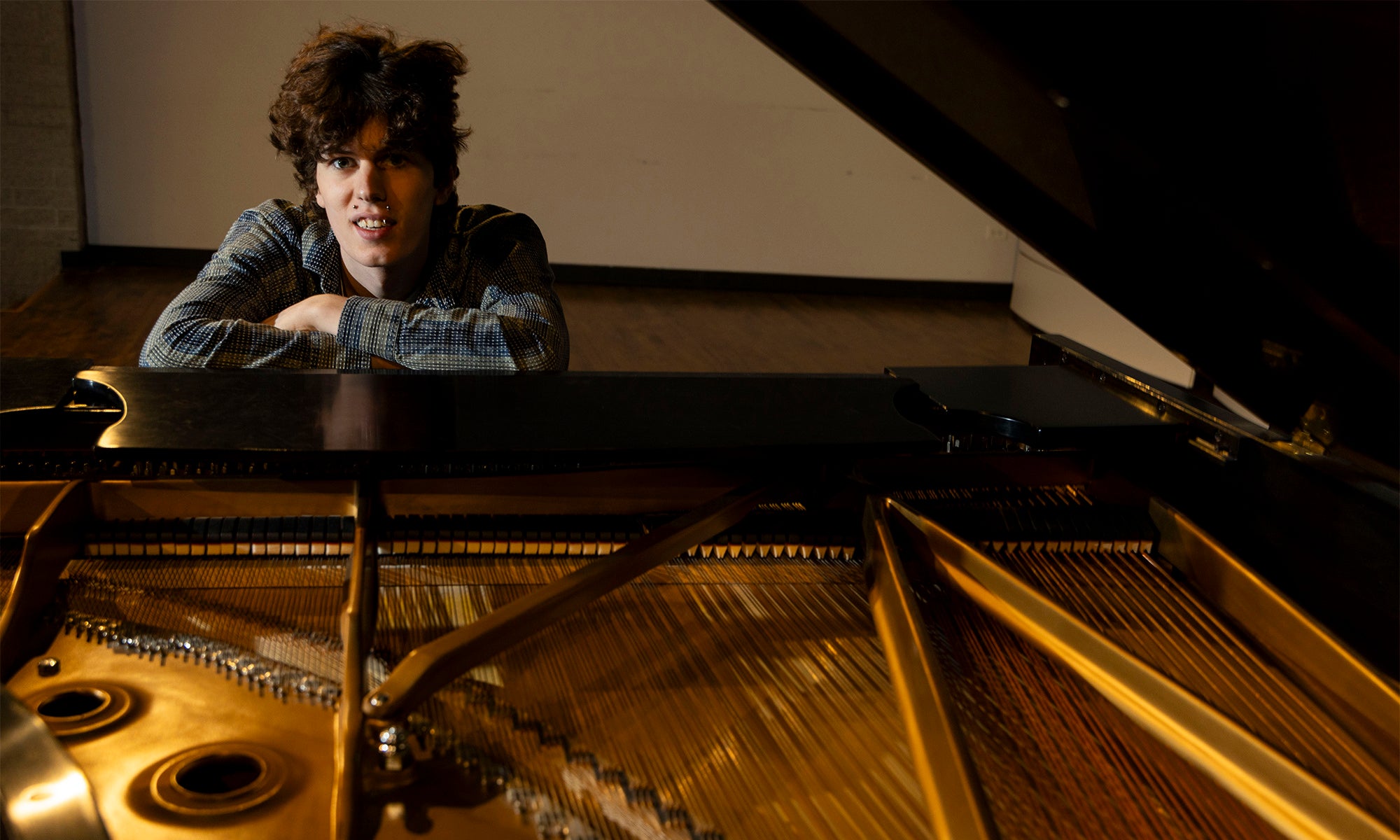 A person with curly hair leans on a grand piano, looking toward the camera. The pianos strings and internal components are visible in the foreground.