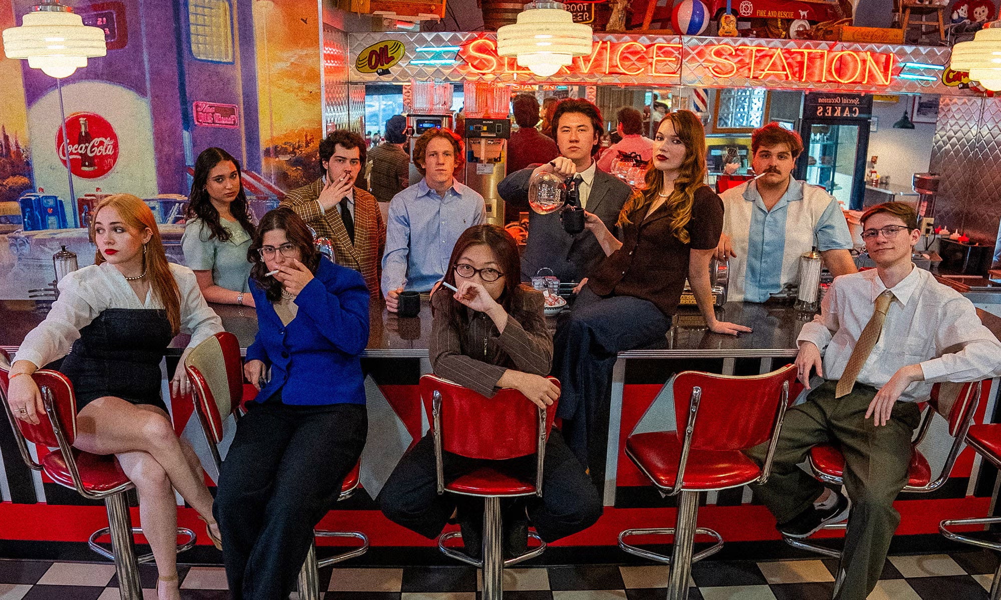 Twelve people in retro-style clothing sit and stand around a diner counter with red bar stools, under bright neon lights and vintage decor.