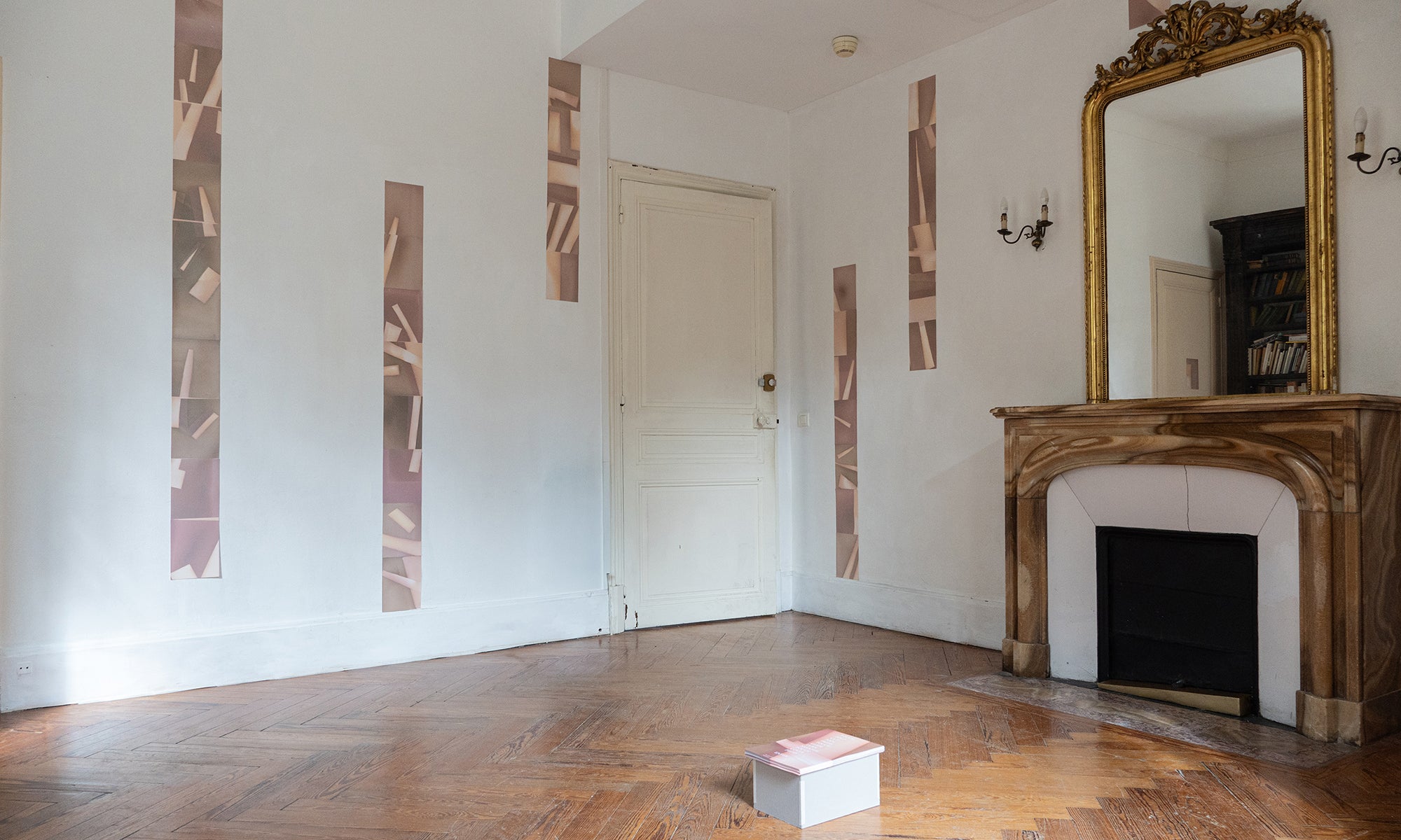 Empty room with a wooden floor, marble fireplace, large ornate mirror, white door, and vertical abstract art on the walls; a closed book lies on the floor.