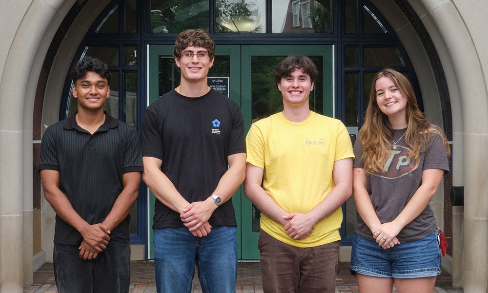 Four young adults stand in a row outside a building with an arched doorway, smiling at the camera.