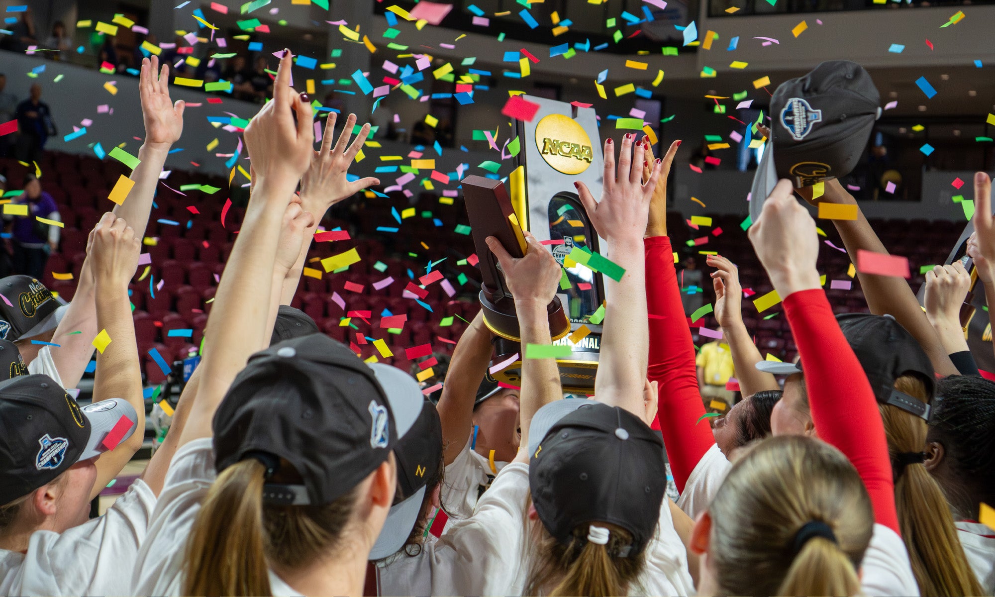 A sports team wearing hats raises a trophy together amid falling confetti in an indoor arena, celebrating a championship victory.