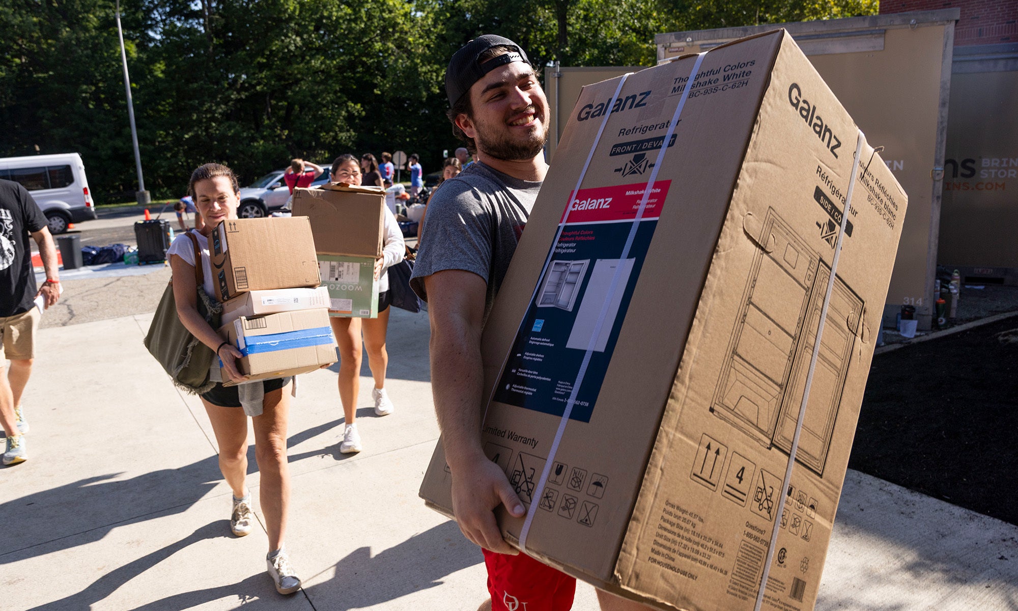 People carry large boxes and belongings outdoors on a sunny day, possibly moving into a dorm or apartment.