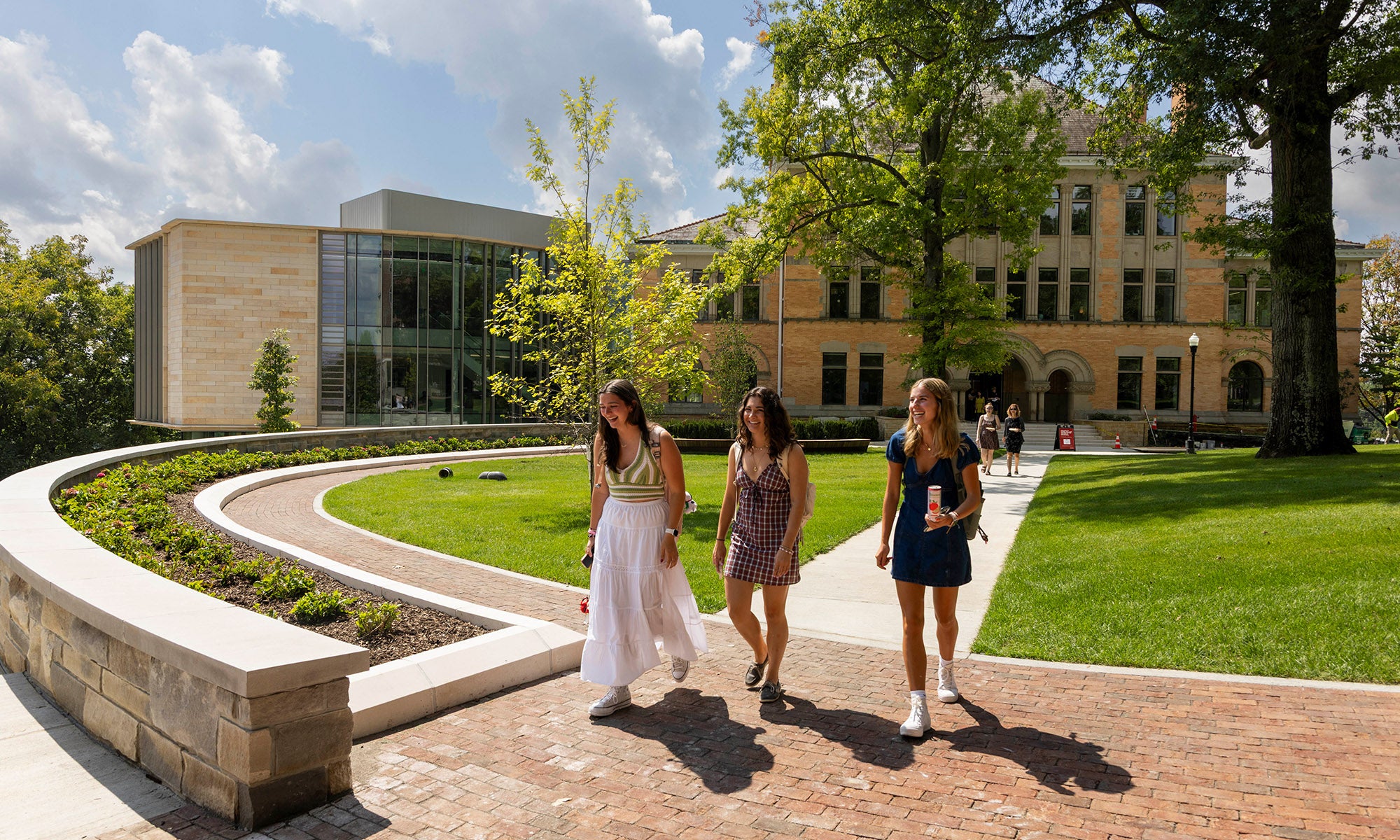 Three people walk along a brick path in front of a modern academic building on a sunny day, surrounded by green lawns and trees.