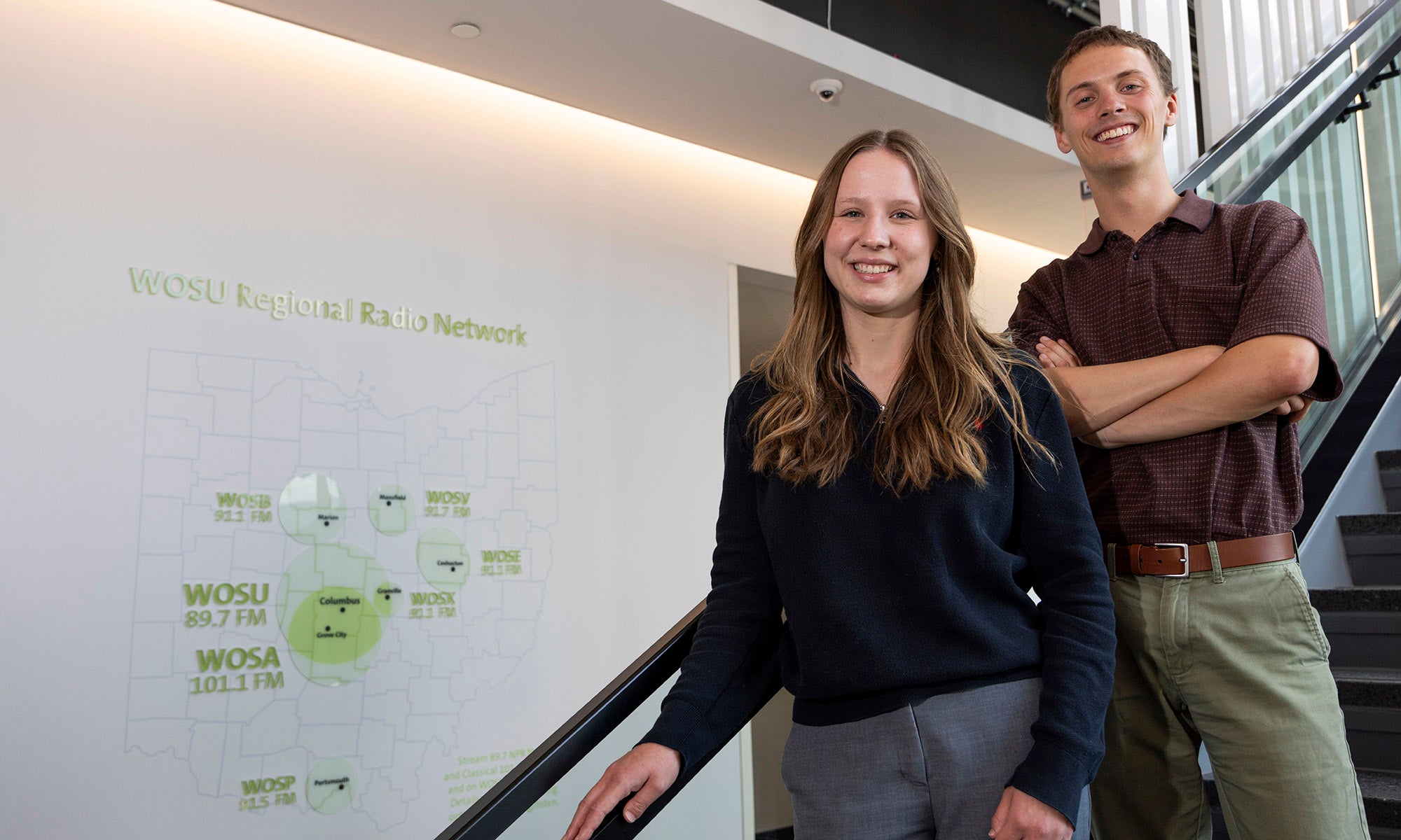 Two people stand side by side on a staircase indoors, smiling at the camera. A diagram is visible on the white wall behind them.
