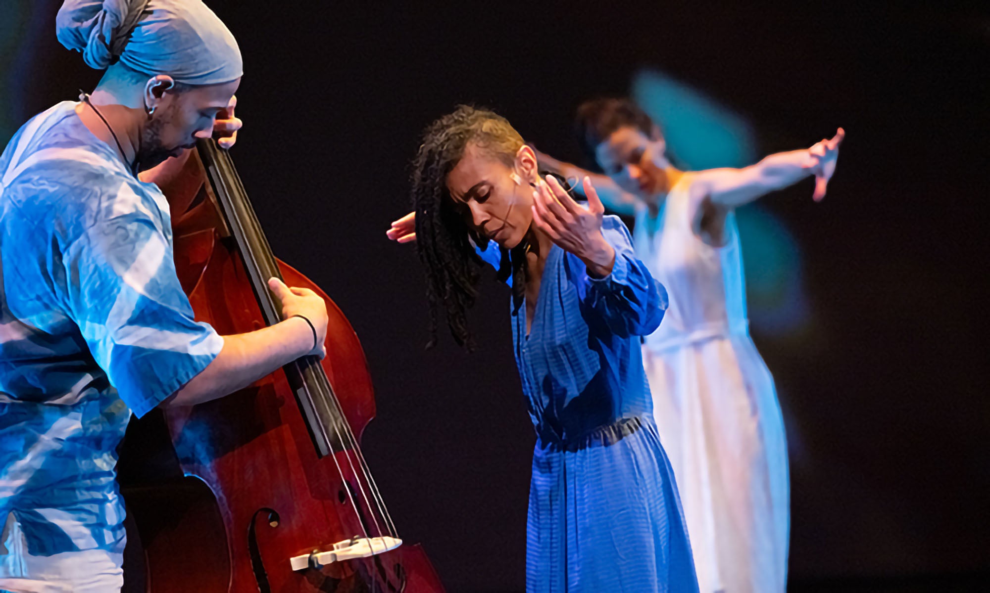 A musician plays an upright bass while two dancers, one in blue and one in white, perform expressive movements on a dimly lit stage.