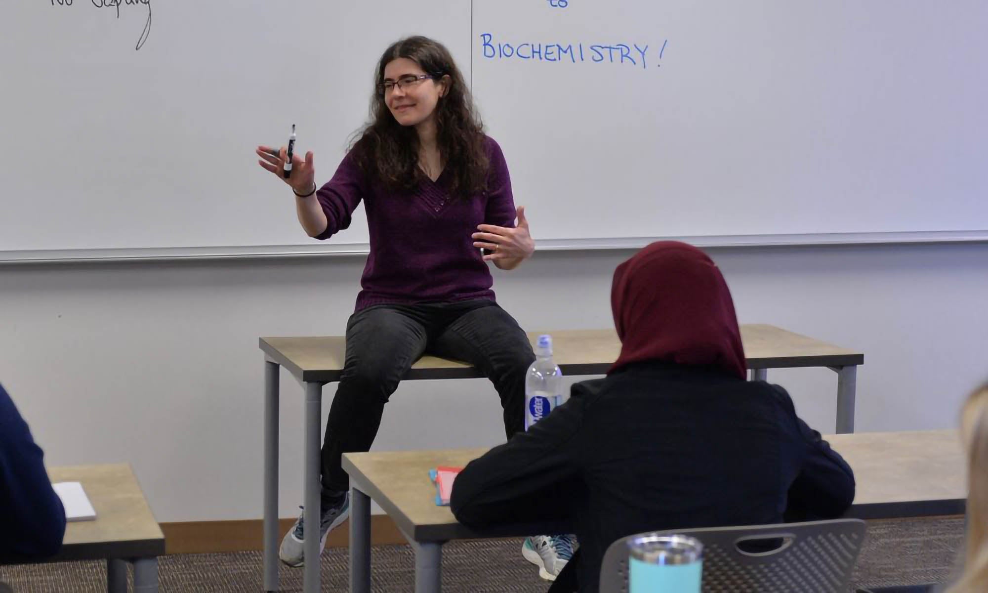 A faculty member sits on a small table at the front of a class of students.
