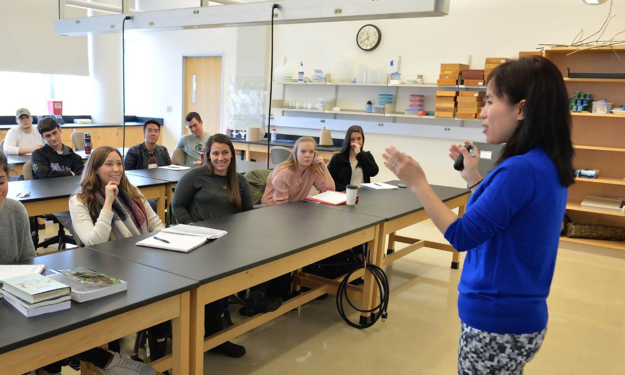 A faculty member in a blue sweater addresses a lab classroom filled with students.