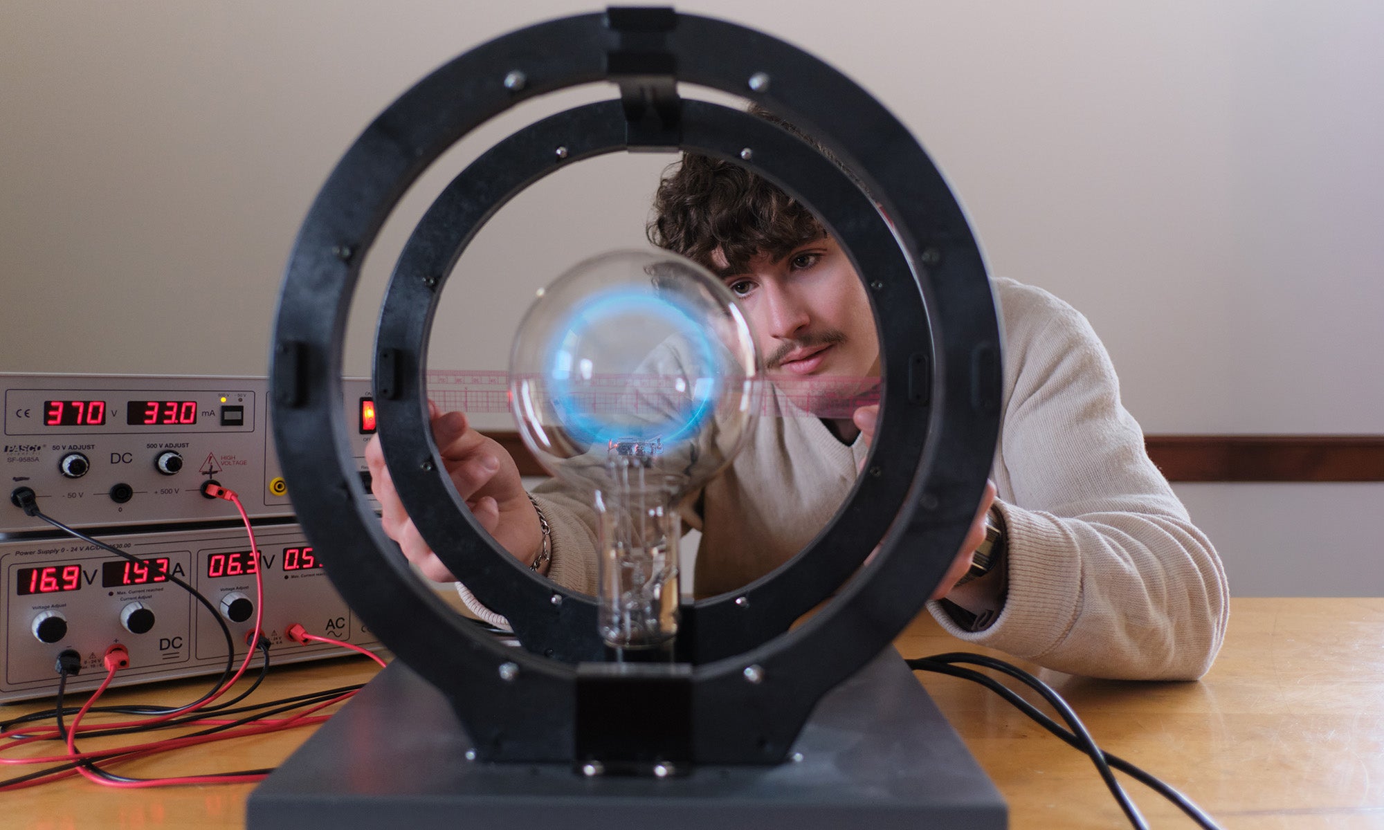 A person adjusts scientific equipment with a glass sphere and magnetic coils, with voltage and current meters in the background on a wooden table.