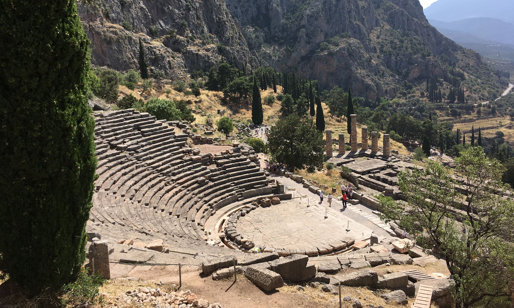 Ancient stone amphitheater with tiered seating is surrounded by trees and mountains under sunlight. A few people stand near the stage area. Columns and ruins are visible in the background.