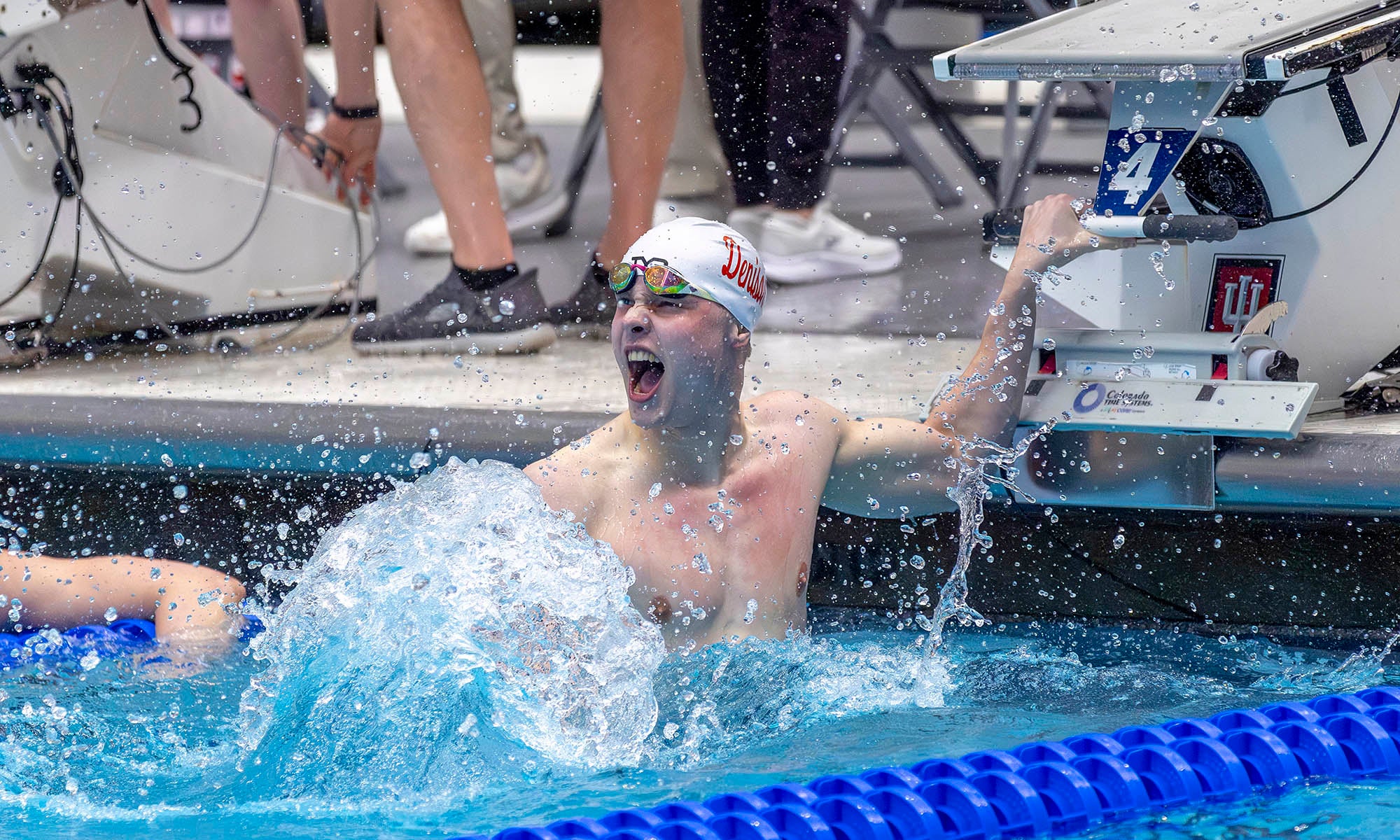 A swimmer wearing goggles and a swim cap is in a pool, splashing water and shouting with an excited expression. People with sneakers are seen in the background near the poolside.