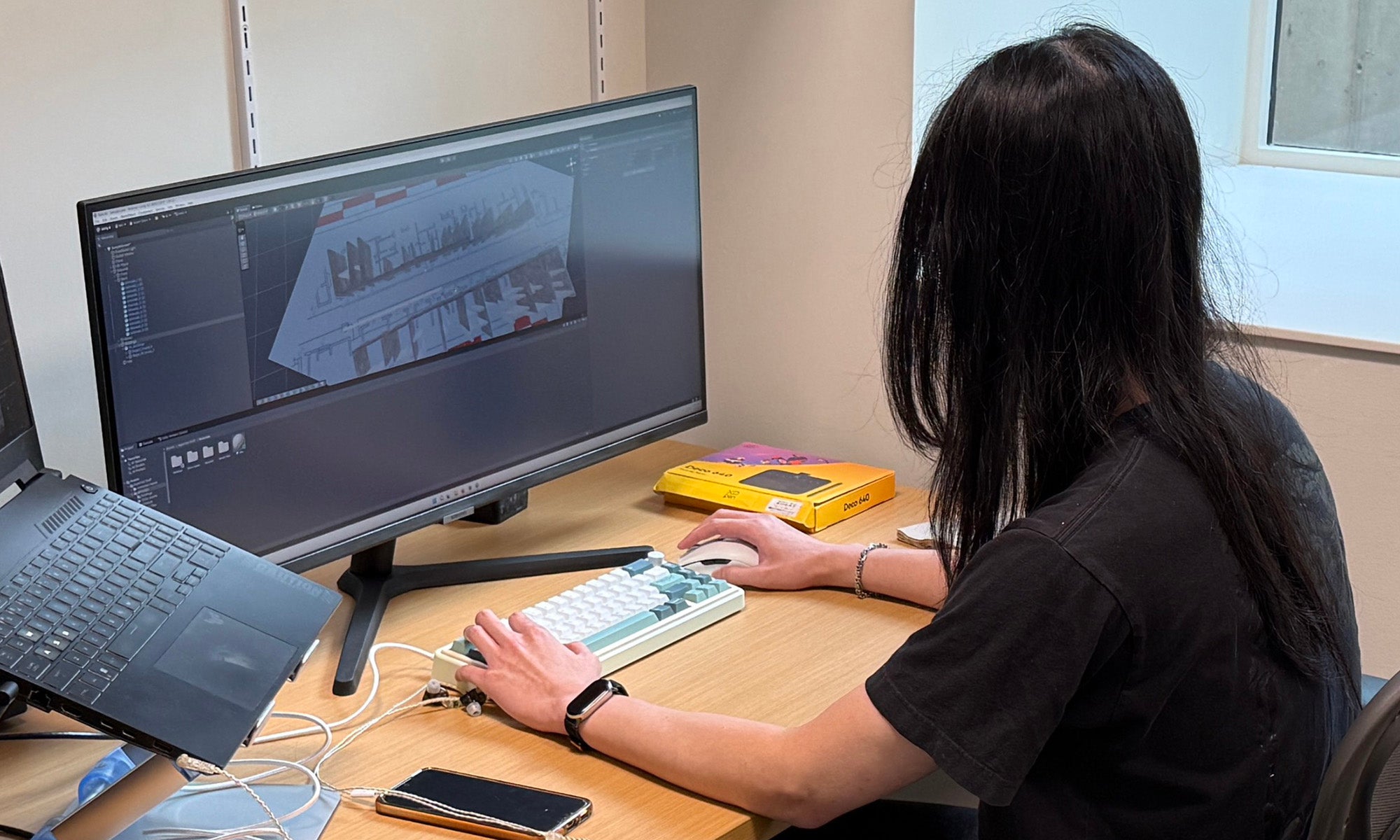Person with long hair sits at a desk using a computer to work on a 3D modeling project; a laptop, phone, and yellow box are also on the desk.