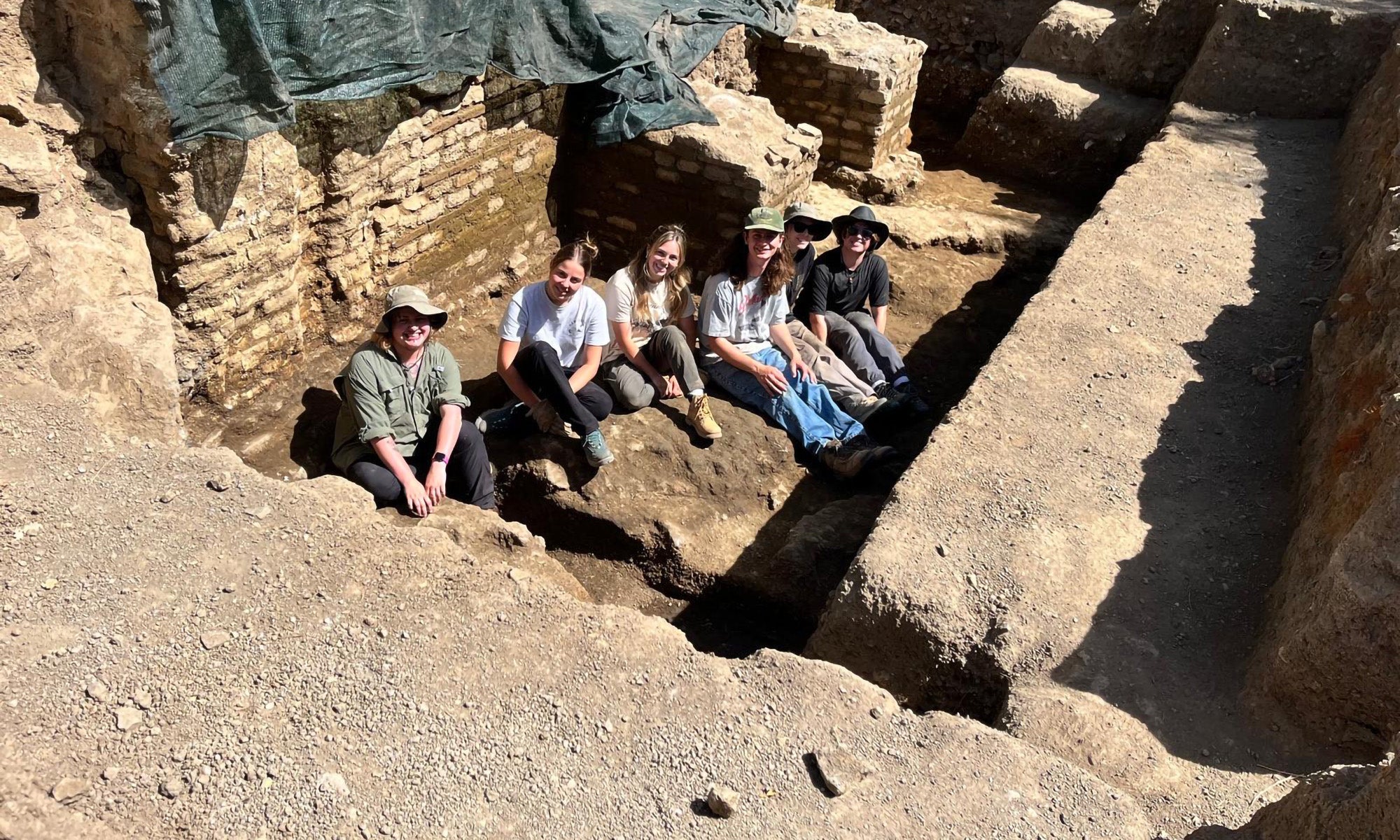 Six people sit in an excavated trench at an archaeological site, surrounded by exposed stone walls and earth, wearing casual clothing and hats.