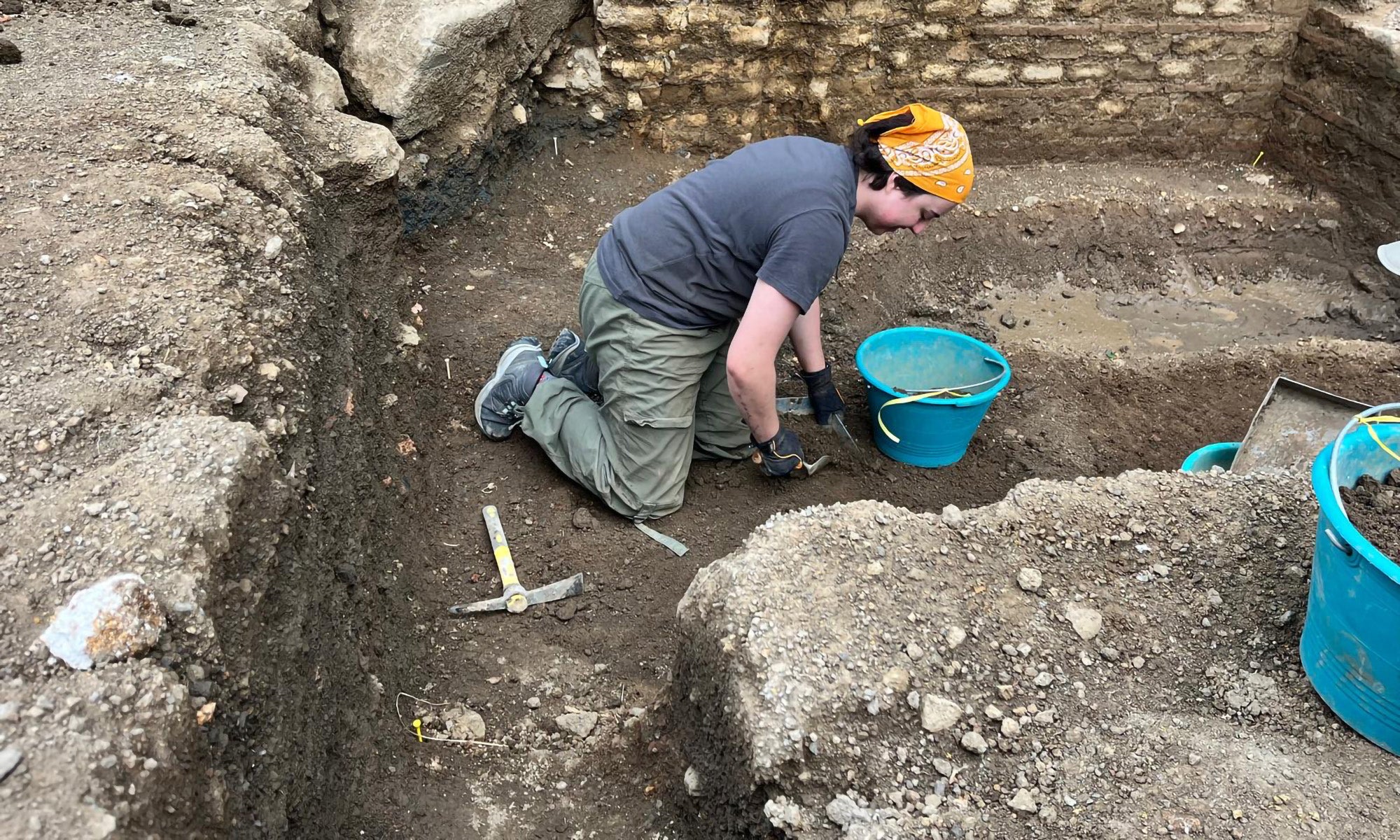 A person wearing a bandana and gloves kneels in an archaeological dig site, using a tool and collecting soil in a blue bucket.