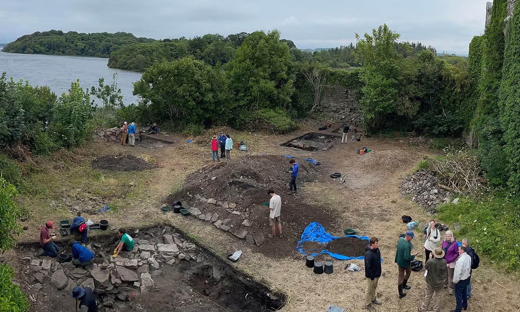 People are excavating an archaeological site near a body of water and old, ivy-covered stone ruins, surrounded by trees under a cloudy sky.