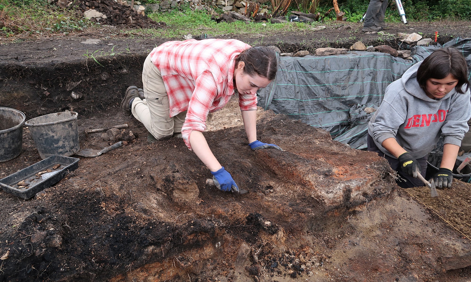 Archaeologists work at a dig site with exposed stone structures, soil piles, and tools, surrounded by trees and a body of water in the background.