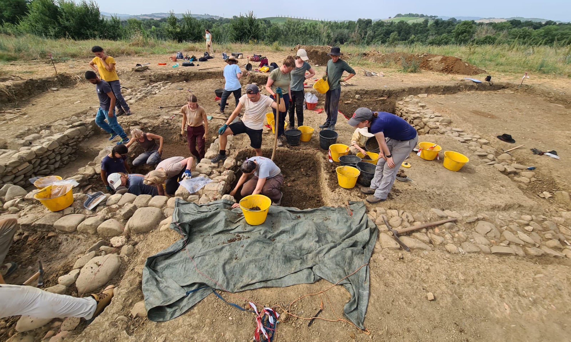 A group of people participate in an archaeological dig, excavating and sorting soil in designated trenches with yellow buckets at a site in an open field.