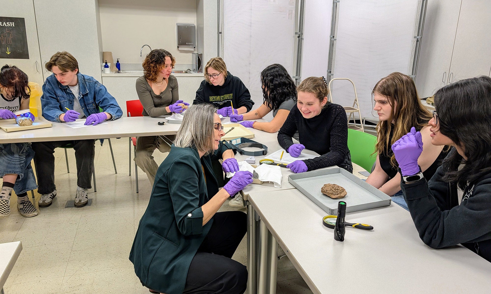 A group of students wearing purple gloves sits around tables, examining objects with a teacher in a classroom setting.