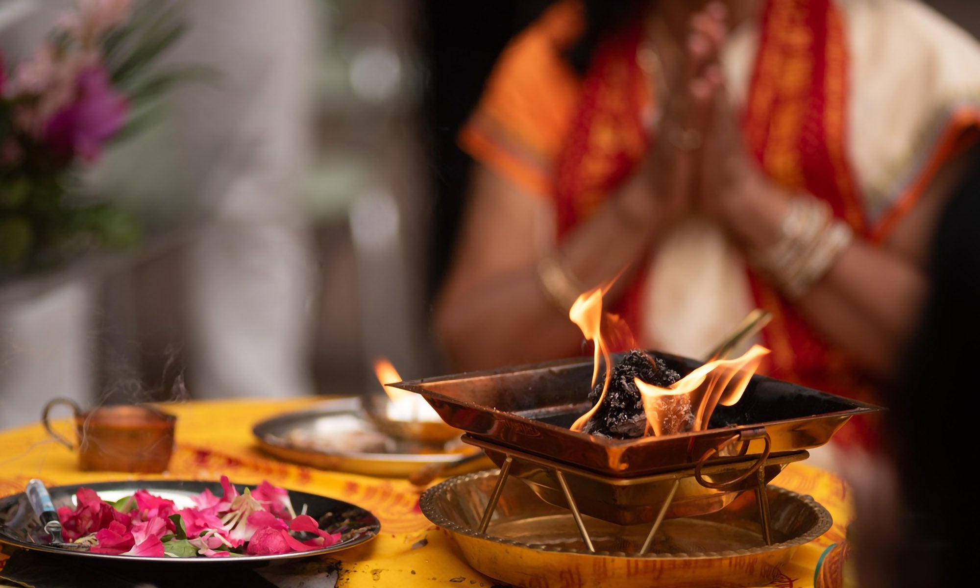 A person in traditional attire sits with hands in prayer near a lit havan kund, flowers, and ritual items on a decorated table.