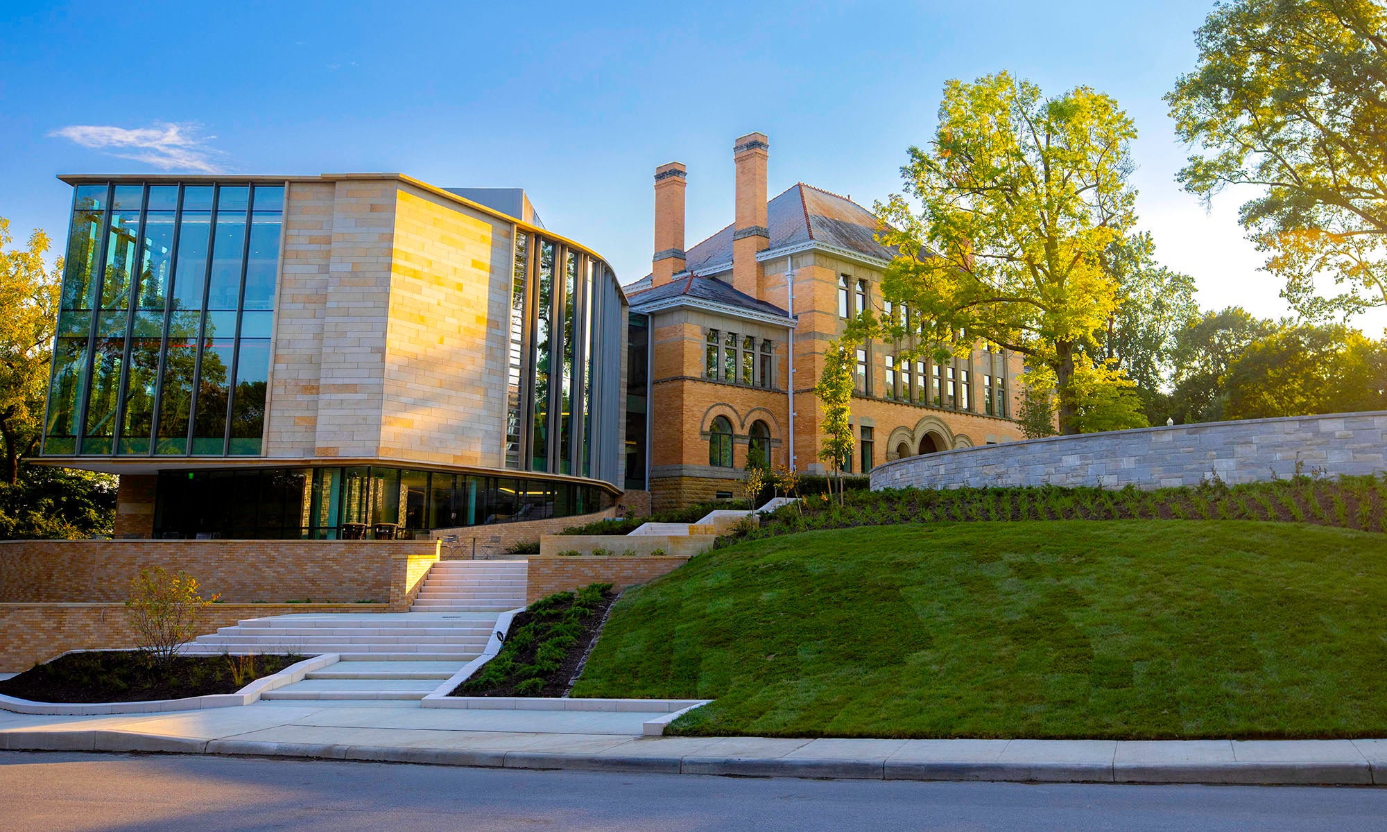 A modern glass extension connects to a historic brick building on a landscaped hill, under a clear blue sky with trees in the background.