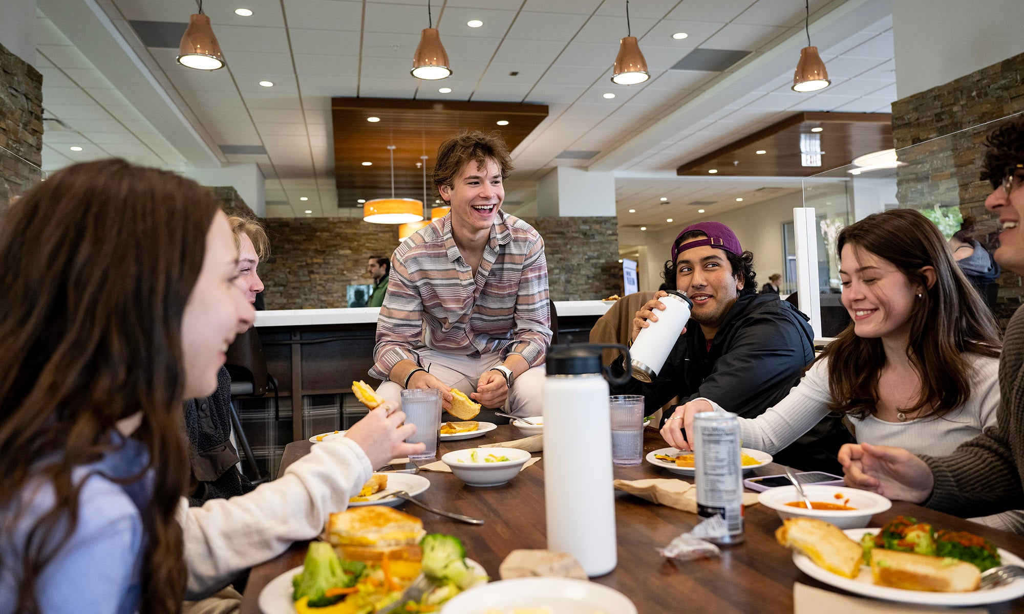 A group of Denison students sit around a table in a dining hall, eating and laughing together. One person sits on a nearby stool, smiling, while others are seated around the table with plates of food and drinks.