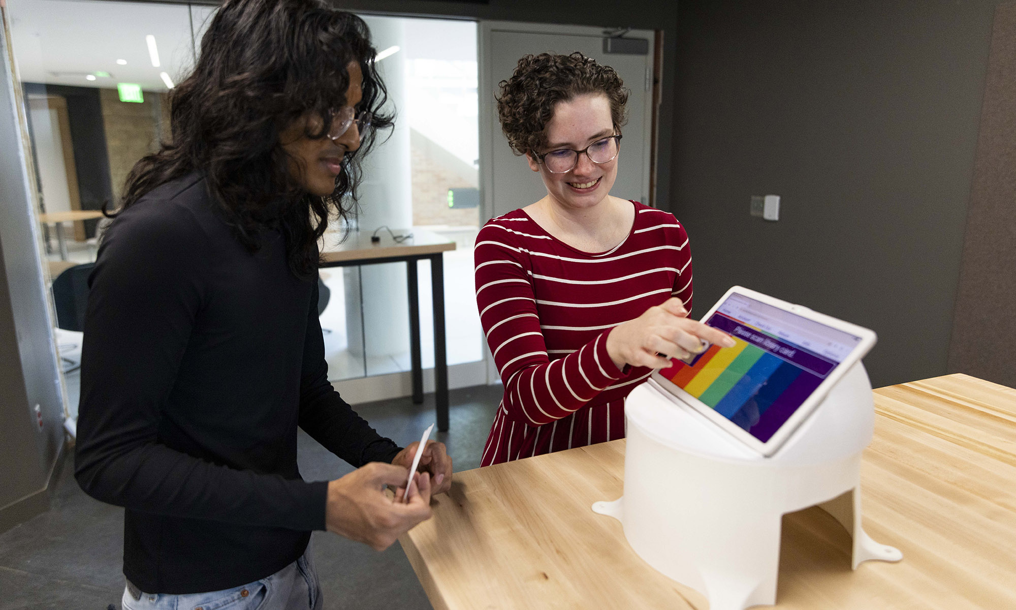 Two people stand at a table while one points at a colorful chart displayed on a tablet propped up on a white stand.