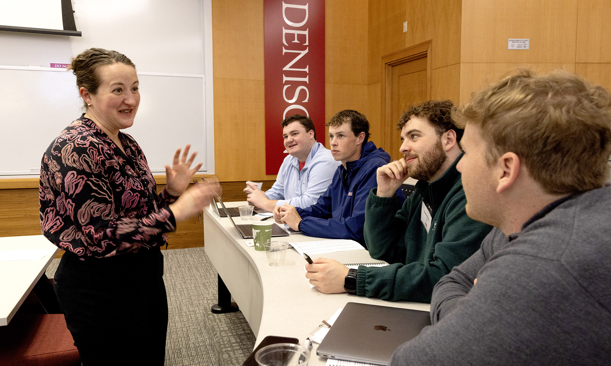A woman speaks to four seated men in a classroom setting with laptops and notebooks on desks; a Denison banner hangs on the wall.