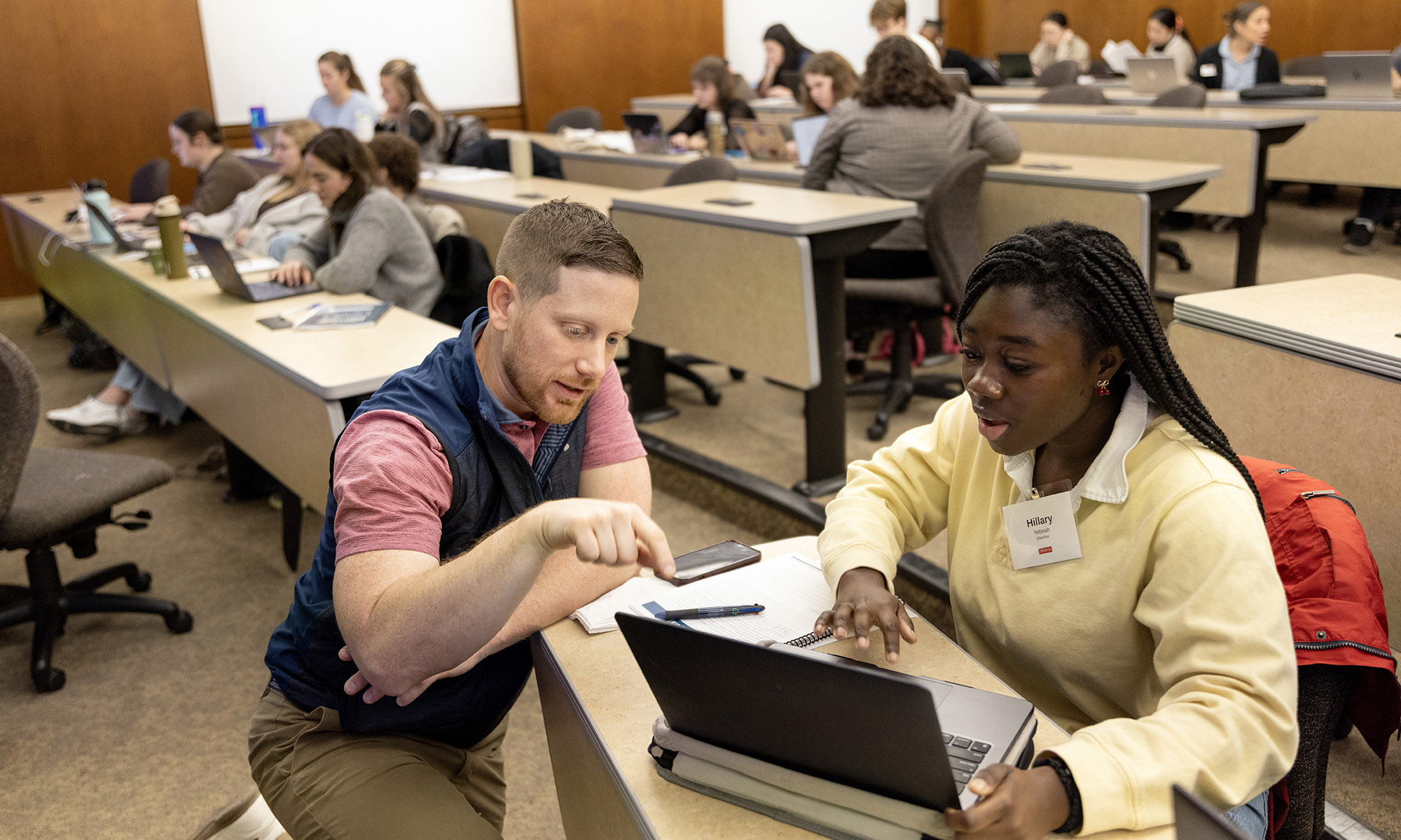A man kneels next to a seated student, pointing at her laptop screen, while others work in groups in a classroom setting.