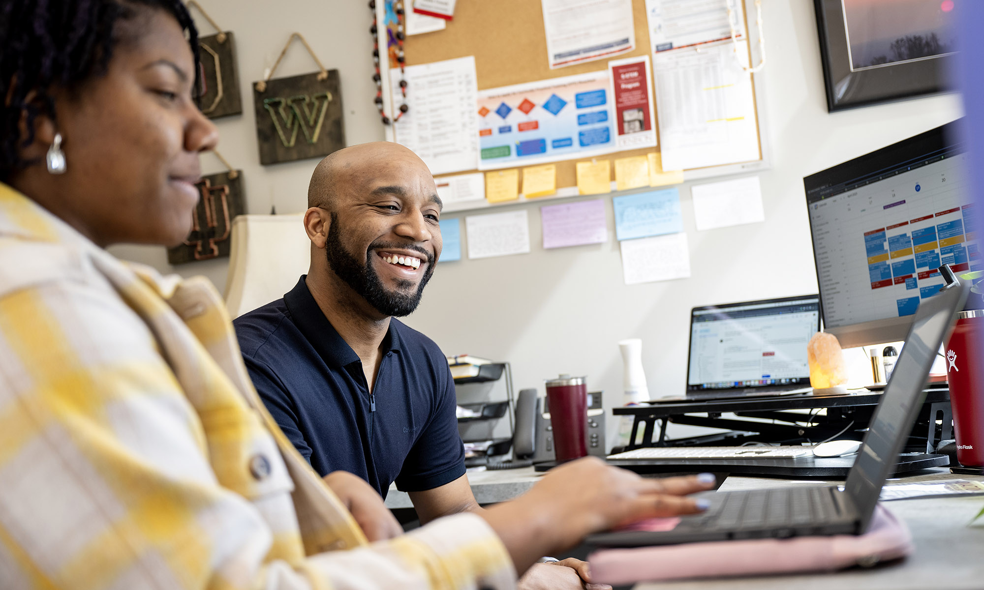 Two people sit at a desk, smiling while working on laptops, with computer monitors, papers, and office supplies in the background.