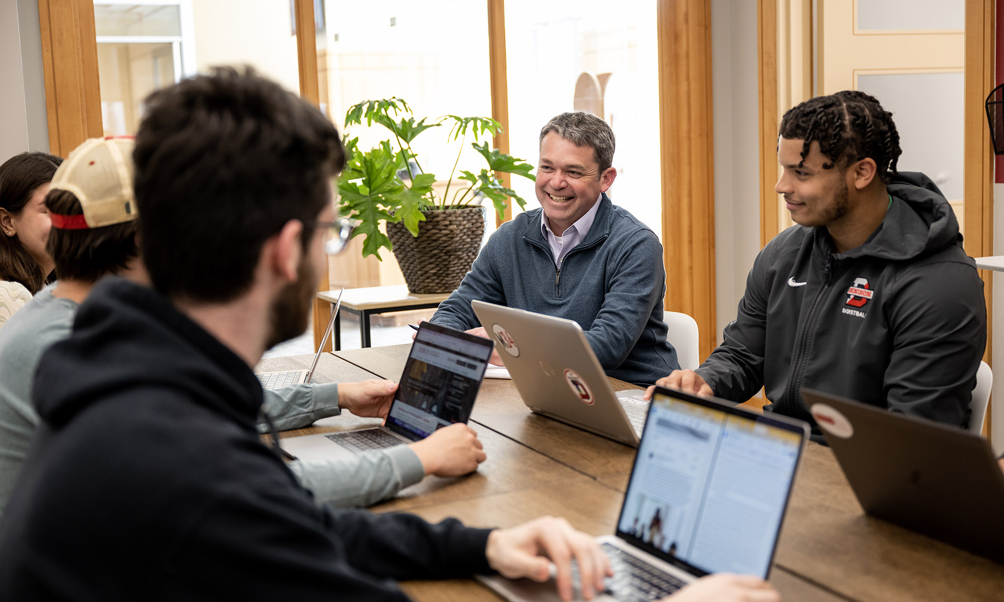 Five people sit around a wooden table with laptops, engaged in discussion. A man in a gray sweater smiles while others listen. Bright room with large plants in the background.