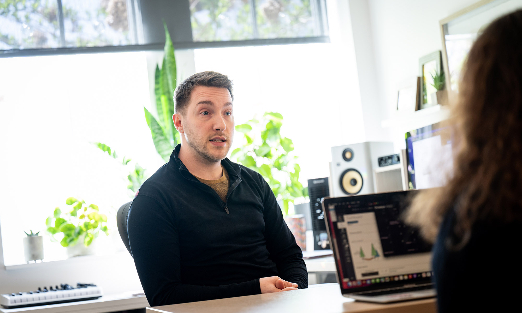 Five people sit around a wooden table with laptops, engaged in discussion. A man in a gray sweater smiles while others listen. Bright room with large plants in the background.
