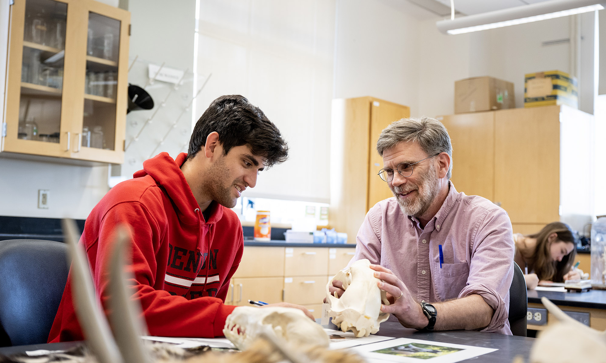 A student and a teacher examine an animal skull together at a table in a science classroom, with study materials and other students in the background.