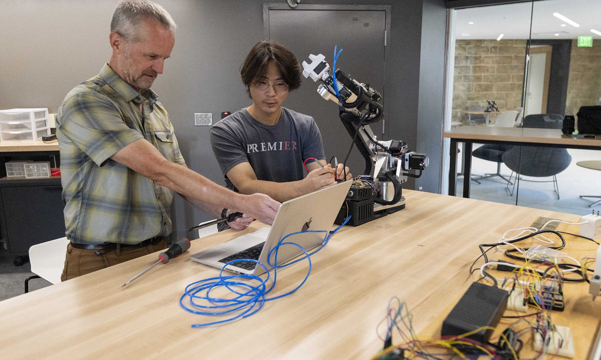 Two people work together at a table with a laptop, cables, and electronic equipment, testing a robotic arm in a modern lab setting.