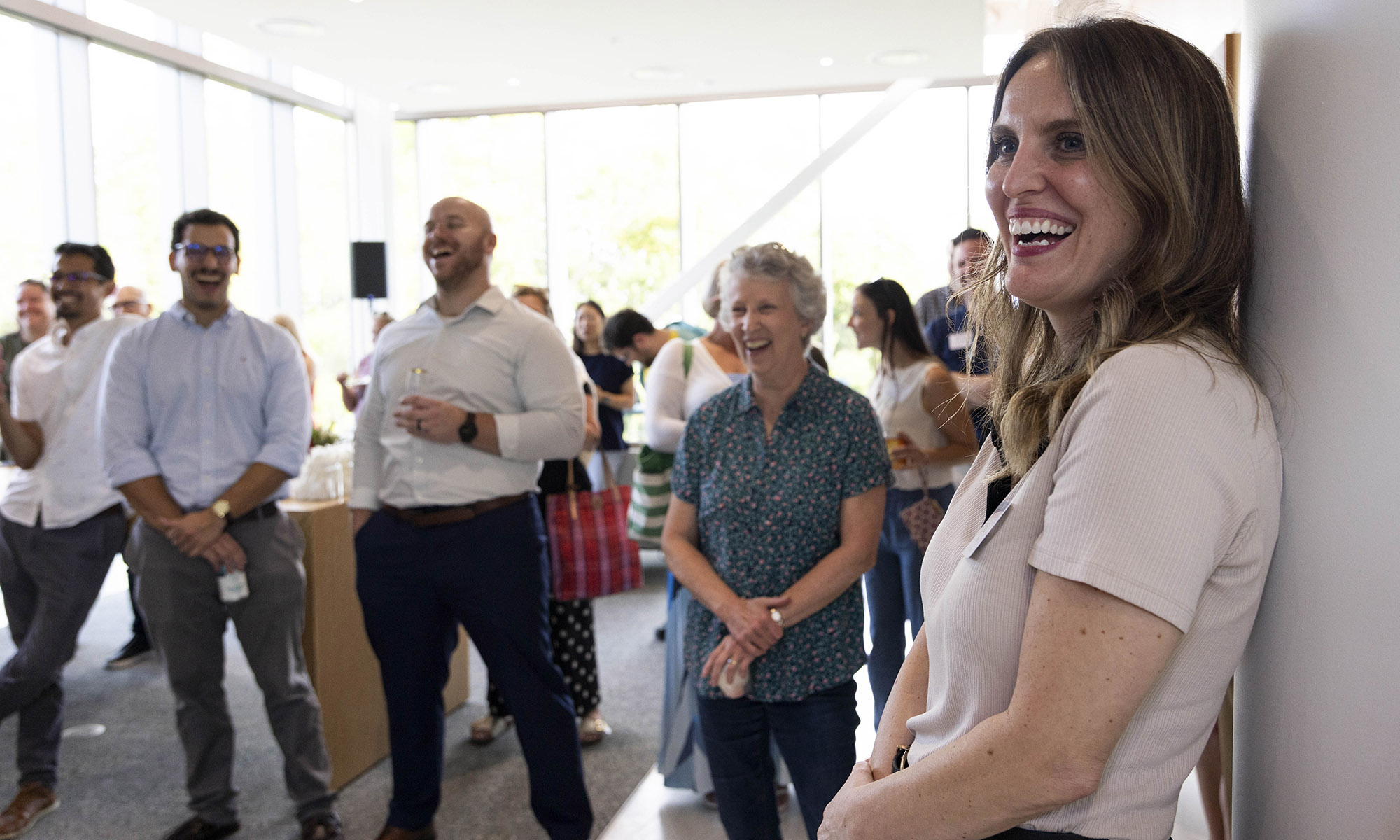 A group of people stand indoors, smiling and laughing during a social or work event. One woman in the foreground leans against the wall, also smiling.