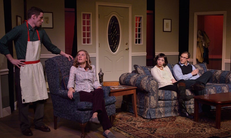 Ken, wearing a feminine apron, leans on the armchair Hannah is seating in. Hannah looks up to him as Beth and John sit on the couch and look at Hannah.Ken, wearing a feminine apron, leans on the armchair Hannah is seating in. Hannah looks up to him as Beth and John sit on the couch and look at Hannah.