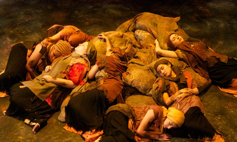 An ensemble of seven Trojan women lie down and sleep together on top of a pile of sacks, lit by a warm yellow light.