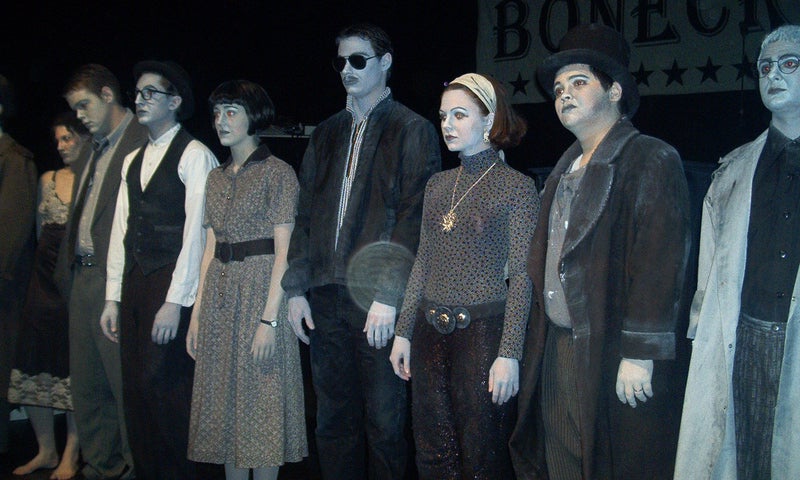 Nine cast members stand in line and face the audience. They are in full greyscale costumes and makeup.