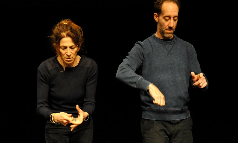 Two actors in dark neutral clothes stand side by side, miming different actions in the piece Marriage: a Work in Progress.