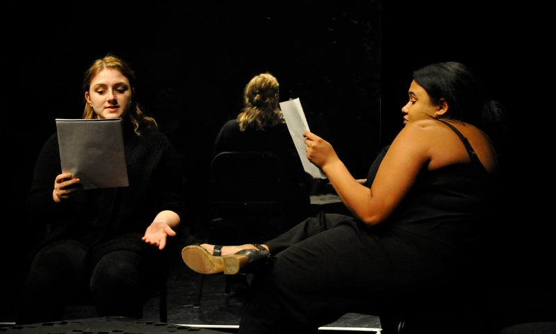 Two actors wearing black sit across from each other, holding scripts as they perform a staged reading.