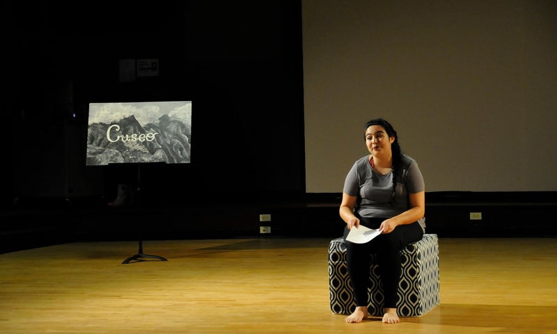 An actor sits on a cube and holds a script as they perform their original piece Esta Soy Yo in the basement of Blair Knapp Hall. To the left of the actor is a small sign on a music stand reading “Cusco” over a painting of mountains.An actor sits on a cube and holds a script as they perform their original piece Esta Soy Yo in the basement of Blair Knapp Hall. To the left of the actor is a small sign on a music stand reading “Cusco” over a painting of mountains.
