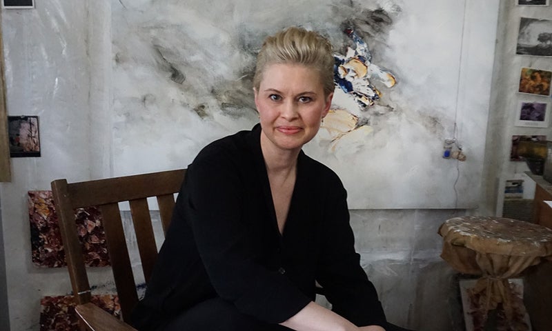 Allison Leigh, a woman with blonde hair, sits on a wooden chair in front of an abstract painting, wearing a black outfit. Artworks and materials are visible in the background.