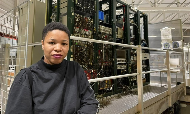 A person stands in front of a large open server rack with exposed internal wiring and equipment.