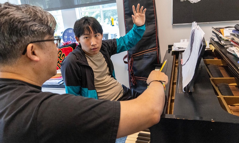  Rumi Kang ’26 sits at a piano discussing his concerto with award-winning composer Professor Ching-chu Hu. 