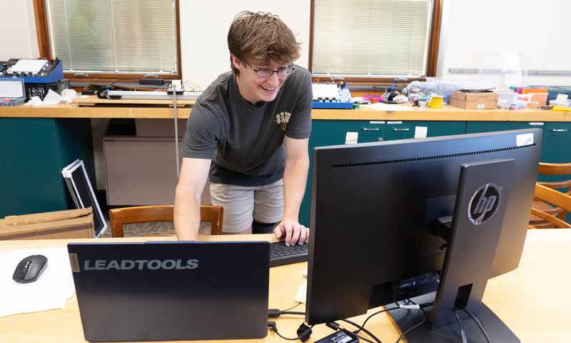 Photo of Denison student Joe Teates ’28 standing at a lab table, looking at computer monitors.