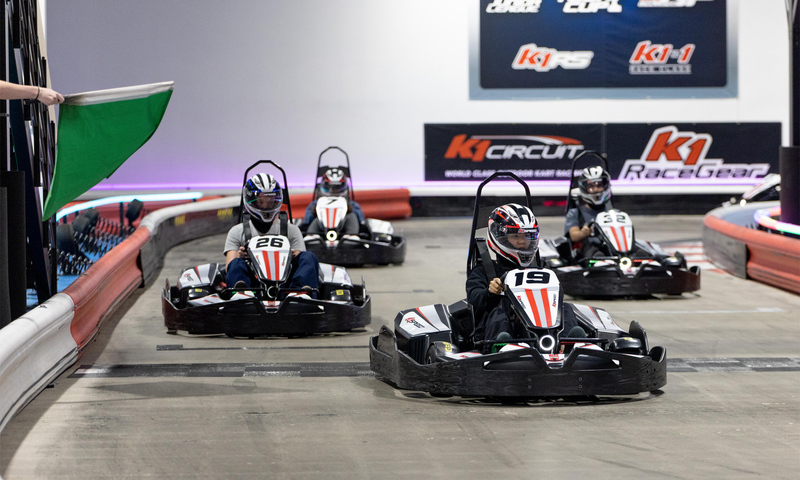 A group of go-karts race on an indoor track as a green flag waves them on.
