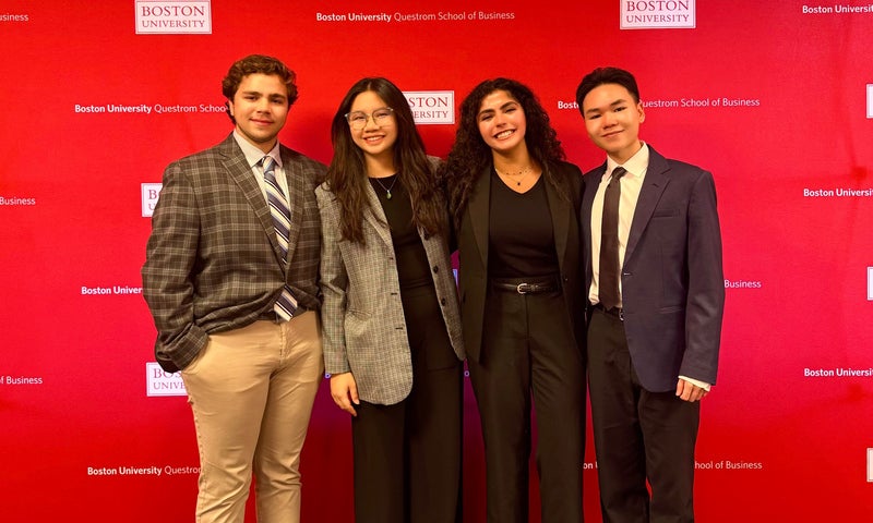 Four Denison students in business attire stand in front of a large red backdrop dotted with Boston University and BU Questrom School of Business logos ahead of their competition.