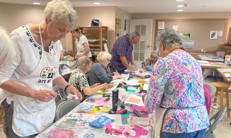 Senior citizens gather around a table filled with art supplies, working together to create art projects.
