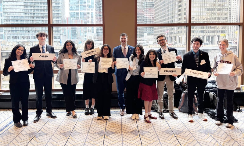 Eleven college students in business attire stand in front of large windows at a conference center, a view of an urban city center behind them, holding certificates and signs displaying the names of countries Oman and Ethopia.