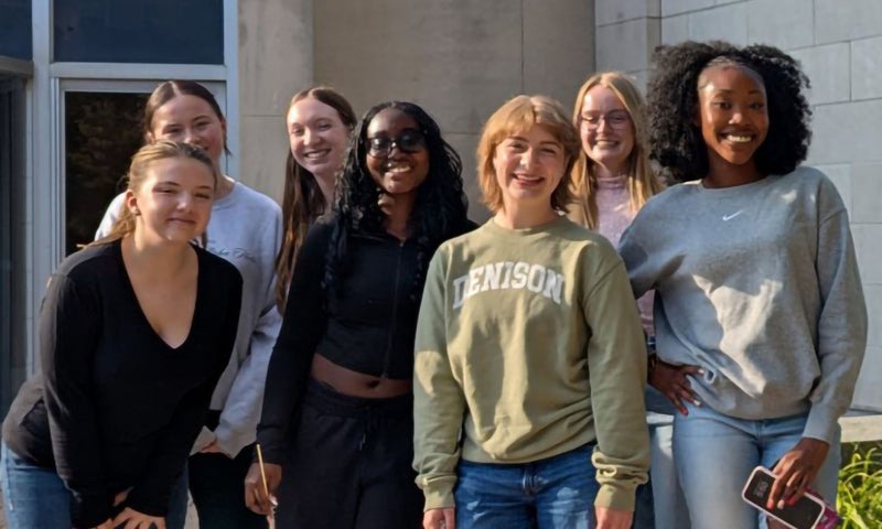 Seven young women stand together outside a building, smiling at the camera. Some wear glasses or sunglasses, and casual clothing.