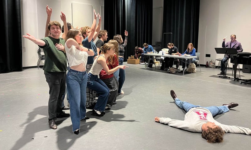 A group of actors rehearsing strike a pose during a choreographed number while the technical team and director watch from the wings.
